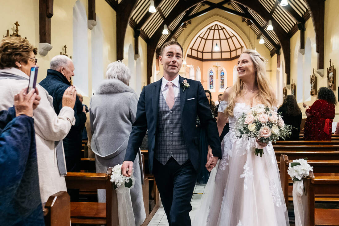 We did it! A photograph of the Bride and Groom leaving the chapel following their nupitals - she is looking at him with a huge smile on her face while they walk together holding hands & watched by family members still in the pews.
