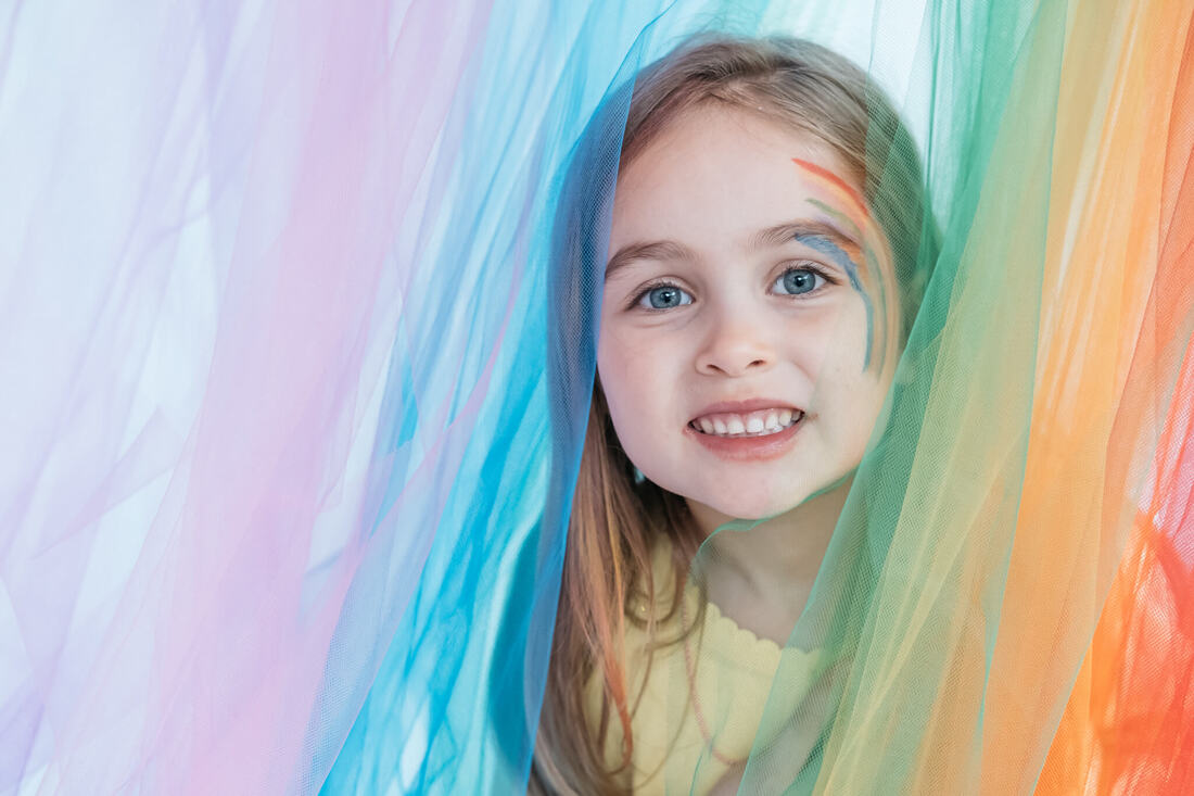 Portrait of a young girl with rainbow face paint looking through rainbow coloured birthday party decorations