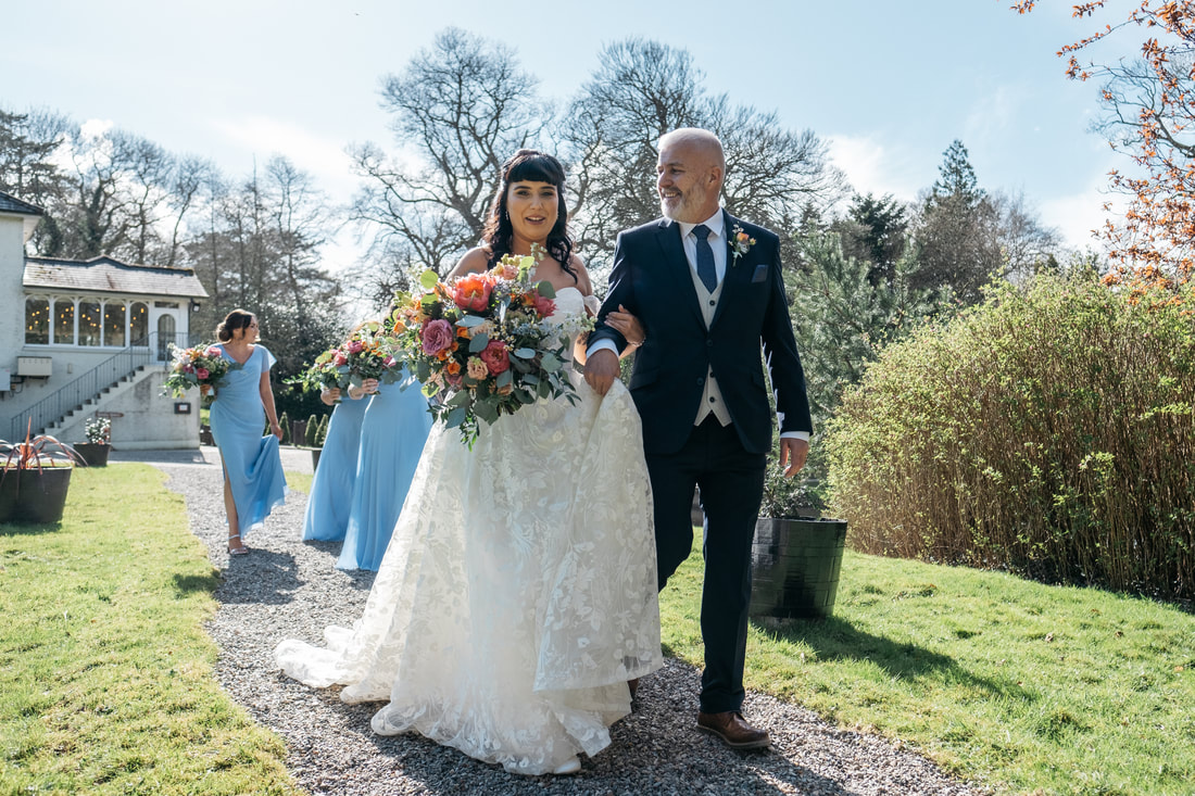 A bride links arms with her dad as they walk through the gardens of the Beech Hill Country House on the way to her Humanist Wedding