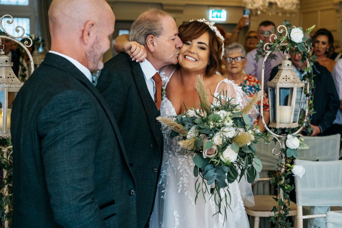 The father of the bride kisses his daughter after walking her up the aisle while the groom and other members of the congregation look on in Bishops Gate Hotel Derry