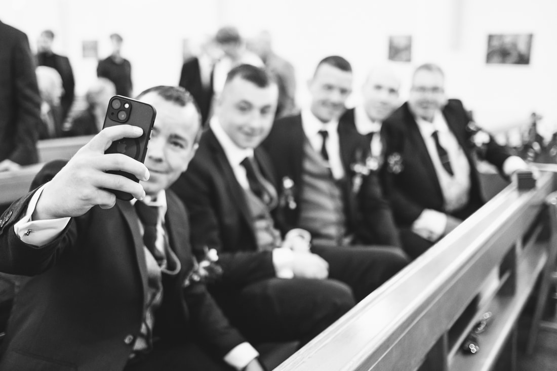 The groomsman closest to the camera takes a selfie with the rest of the groomsman and groom in the front row of the chapel