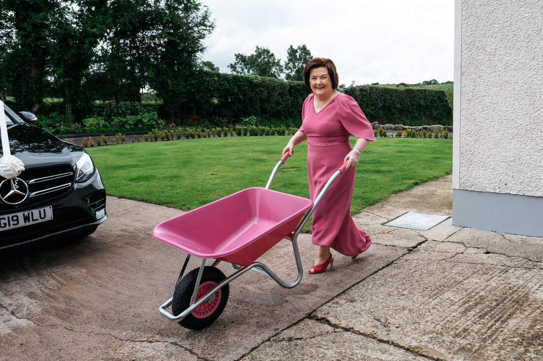 The brides mum pushing a pink wheelbarrow across the garden, while wearing a matching pink dress on the morning of a Tyrone wedding