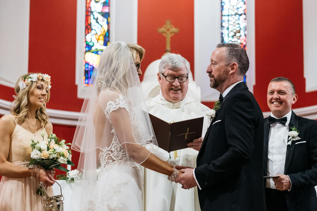 a photograph of the bride and groom holding hands while they stand in front of the priest making their wedding vows - while the Best Man & Matron of Honour watch on