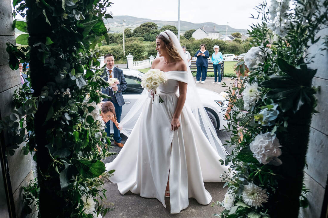 A photograph of a bride framed by flowers as she is prepared on the steps of the chapel before she walks up the aisle at the start of her wedding in County Donegal
