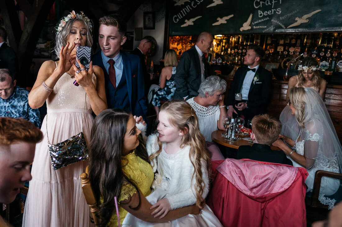A colourful photograph of a busy bar scene during a wedding day - at the front of the image a bridesmaid is taking a selfie on her phone - the flower girl is sitting on a family friends lap - the bride and page boy are at a table behind them while others talk all around the image 