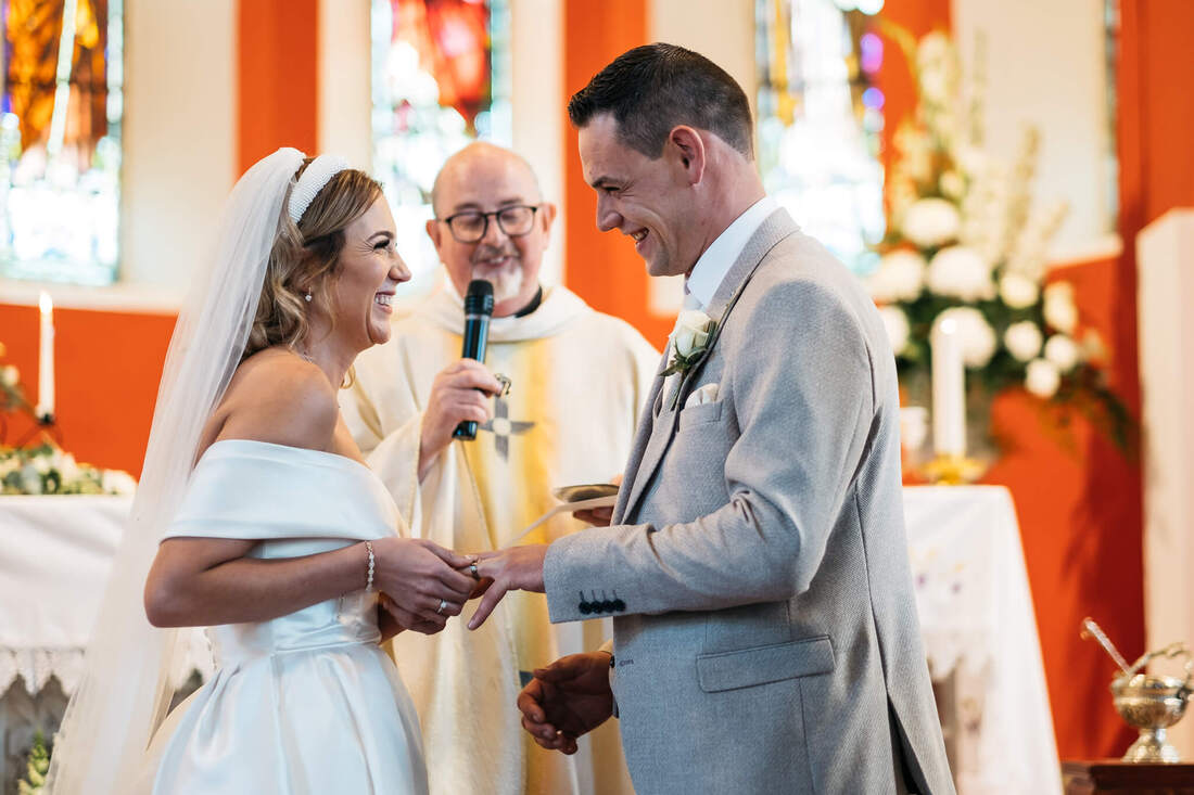a photograph of the bride and groom sharing a moment of laughter on the alter as she places the wedding ring on her new husbands finger in front of the priest during their Donegal wedding