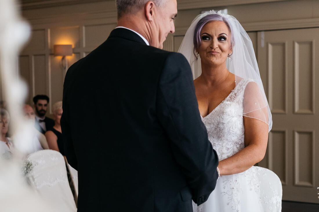A photograph of the bride giving a flirtatious look to her groom during their wedding vows at a civil ceremony in Bishops Gate Hotel Derry