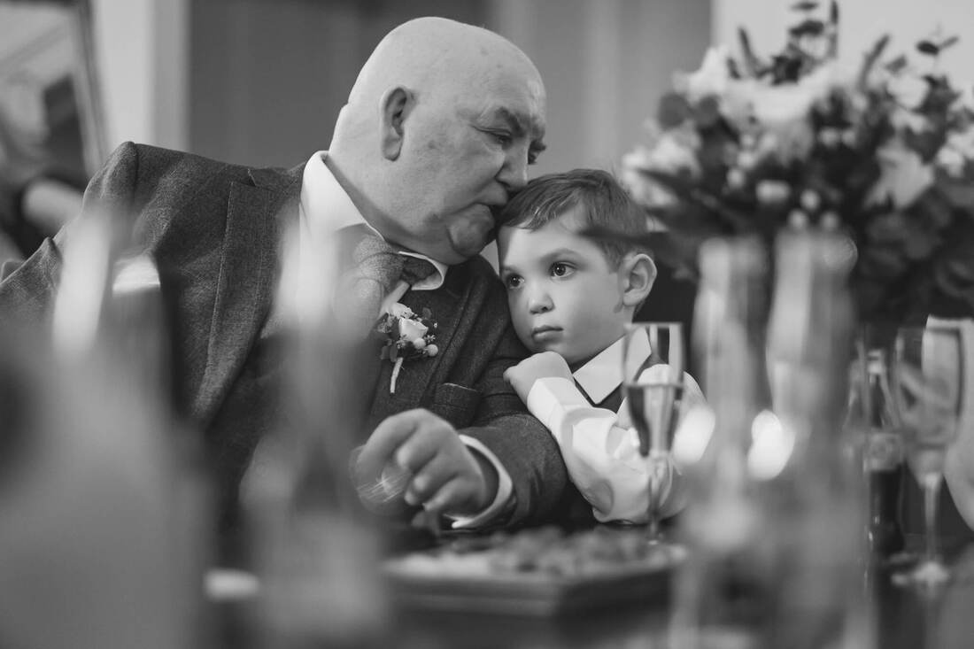 A black and white image of a touching moment as the father of the bride leans to the side and kisses his grandson on the head during a wedding reception in Derry