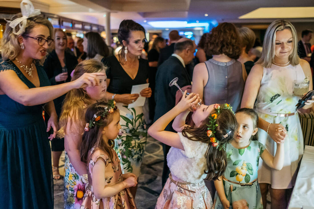 A photograph taken during a wedding reception in Derry - a young flower girl has raised a champagne glass to her mouth and is taking a drink while a nearby adult is putting and trying to get her to stop before she drinks the champagne while other guests look on