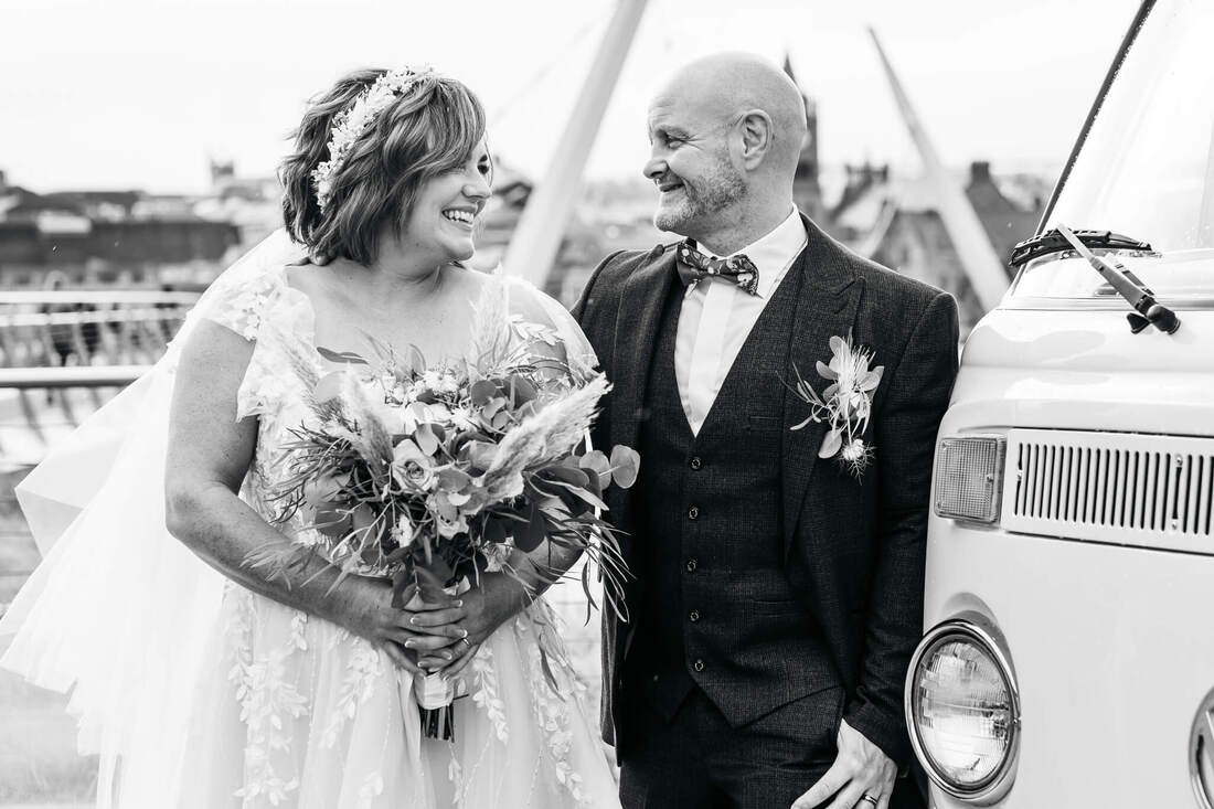 Bride and Groom smiling at each other, beside a VW Van with the Derry Guildhall & Peace Bridge behind them