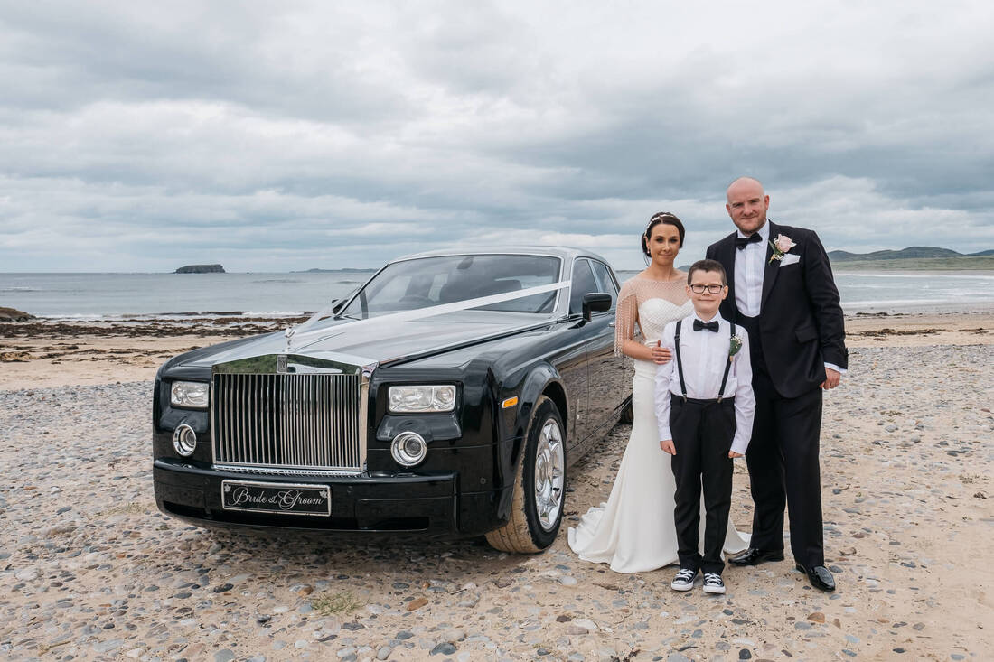 A bride and groom, with their pagesboy son, on the shore of Pollan Bay, Ballyliffen, standing alongside a Rolls Royce on their wedding day