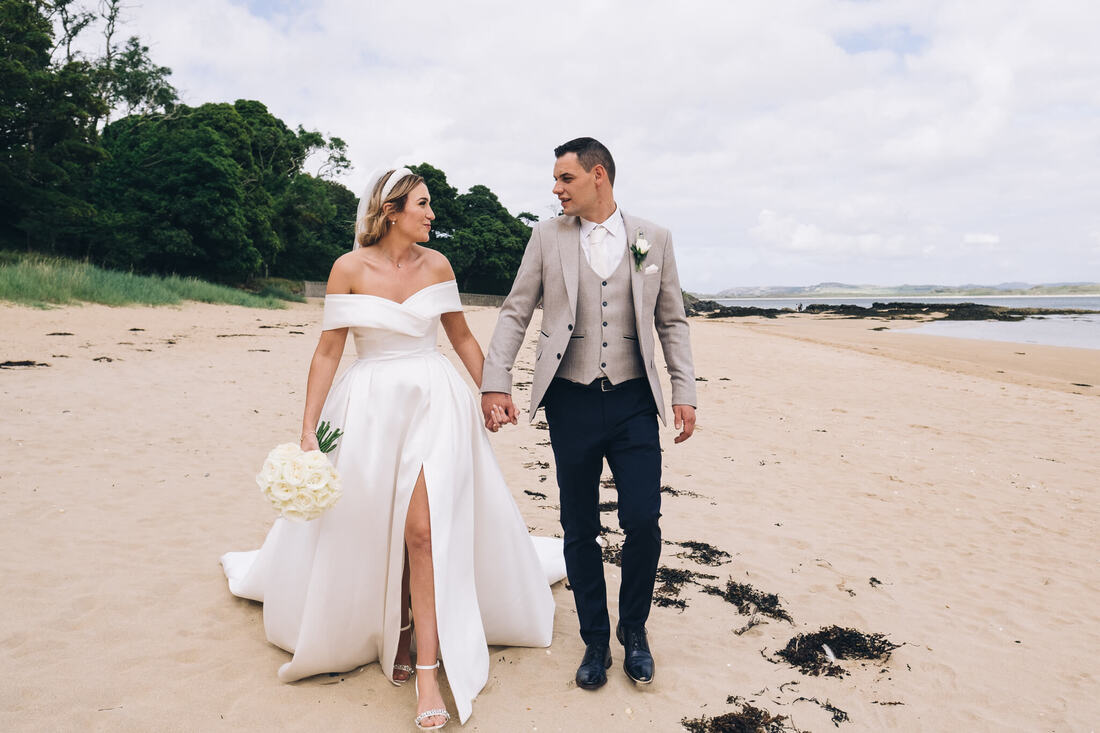A bride and groom walk hand in hand along the shoreline at Ards Friary Beach in County Donegal
