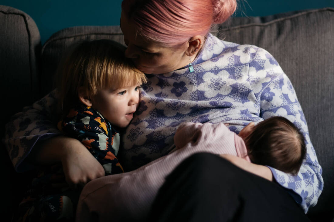 A pink haired mum cuddling her two children - kissing her son on his head, while his baby infant daughter is fed